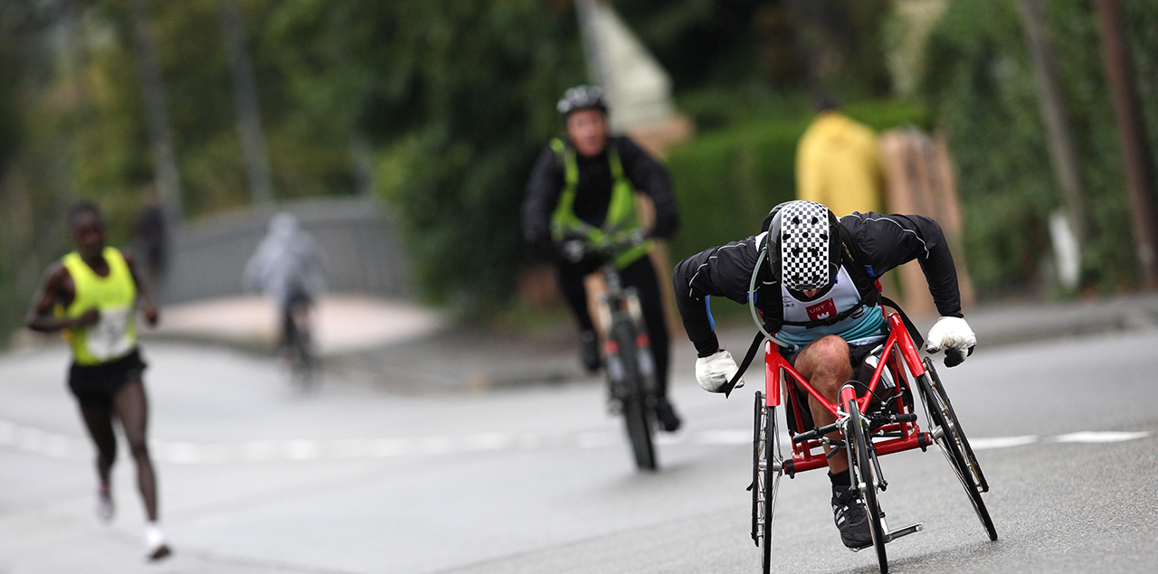 A wheelchair cyclist, a person running and a person riding a bicycle together on an outdoor road