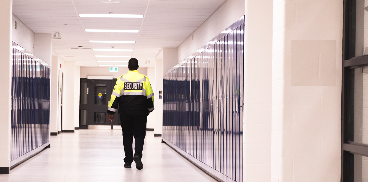 The back view of a security guard walking down an empty school hallway.